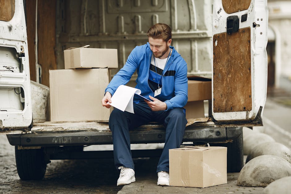 A man seated on the edge of a white delivery van's open back door during a house relocation, surrounded by moving boxes, including cardboard boxes stacked inside the van and on the ground nearby. The man is holding a clipboard and appears to be reviewing inventory or packing details, wearing casual clothing with a blue hoodie and white sneakers. The interior of the van shows shelving and packaging materials such as plastic wrap and blankets, used to protect furniture during transport. The scene takes place outdoors on a paved area, with large rocks visible in the background. The loading process is part of a furniture transport or packing and moving operation, with temporary lighting illuminating the area. This setup is indicative of professional removals services, possibly provided by Epsom Man and Van, who handle home relocation and logistical arrangements.