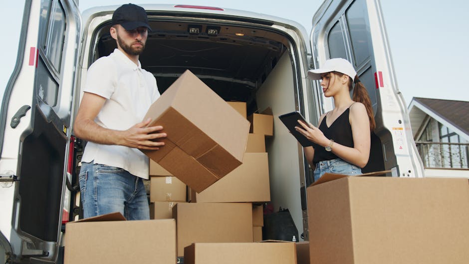 A man and a woman are working together during a home relocation, with the man holding a cardboard box and the woman standing beside the open rear doors of a white van. Inside the van, several stacked cardboard boxes of various sizes are visible, some with packing tape, along with protective materials such as bubble wrap or plastic covering parts of the furniture. The man is wearing a white polo shirt, jeans, and a black cap, while the woman is dressed in a black sleeveless top, jeans, and a white cap, holding a tablet device. The environment appears to be outside a residential property, with a house and clear sky in the background. The loading process involves careful handling of boxes, possibly assisted by trolleys or straps, aligning with a professional removals service like Epsom Man and Van, focusing on packing and furniture transport for a house move.