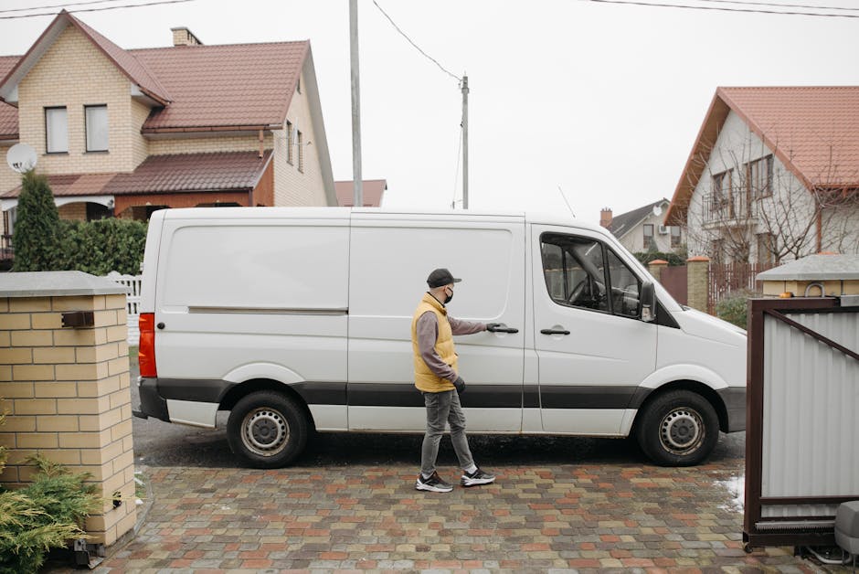 A man seated on the edge of a white delivery van's open back door during a house relocation, surrounded by moving boxes, including cardboard boxes stacked inside the van and on the ground nearby. The man is holding a clipboard and appears to be reviewing inventory or packing details, wearing casual clothing with a blue hoodie and white sneakers. The interior of the van shows shelving and packaging materials such as plastic wrap and blankets, used to protect furniture during transport. The scene takes place outdoors on a paved area, with large rocks visible in the background. The loading process is part of a furniture transport or packing and moving operation, with temporary lighting illuminating the area. This setup is indicative of professional removals services, possibly provided by Epsom Man and Van, who handle home relocation and logistical arrangements.