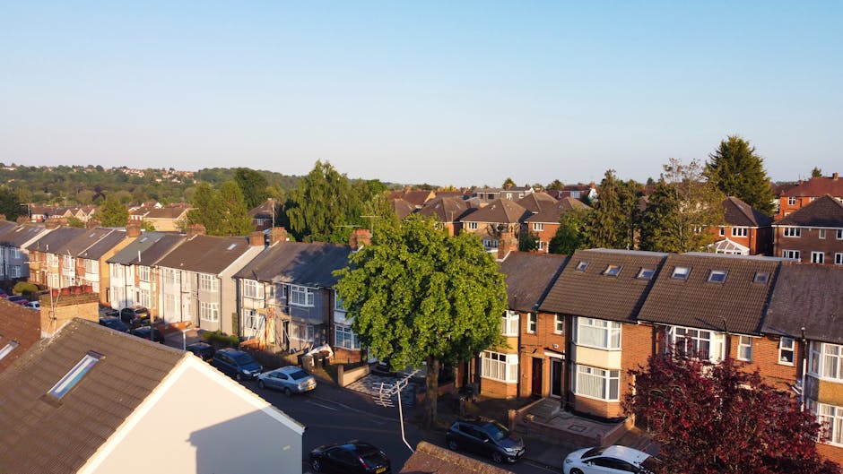 An aerial view of a residential street featuring a row of terraced houses with brick and pastel-colored facades, some with solar panels on the roofs. Cars are parked along the curb, and a large green 