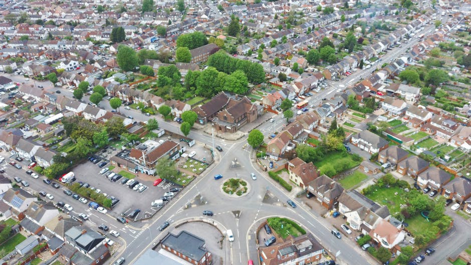 An aerial view of a residential street featuring a row of terraced houses with brick and pastel-colored facades, some with solar panels on the roofs. Cars are parked along the curb, and a large green 