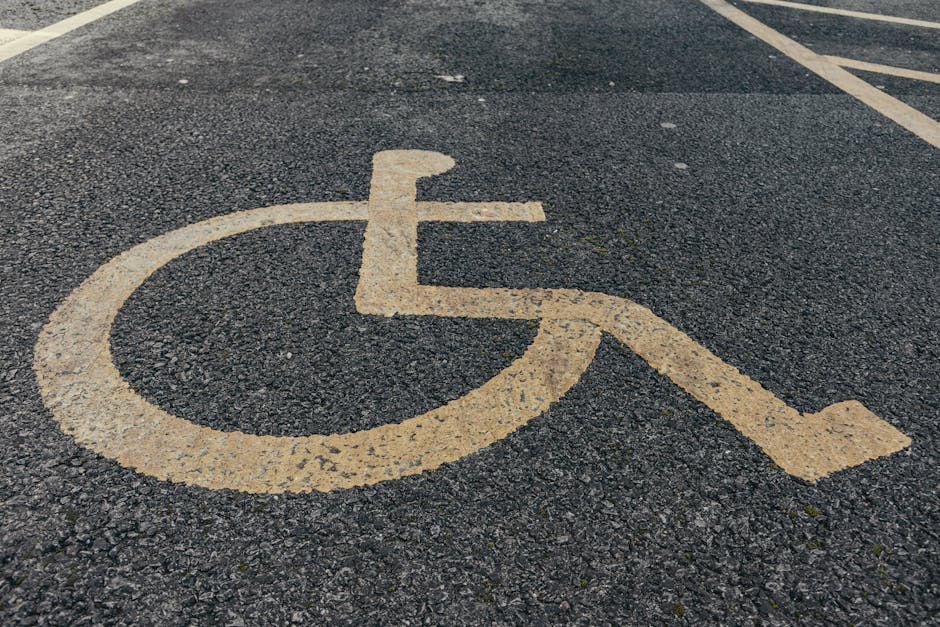 A close-up view of a yellow wheelchair accessibility symbol painted on a dark asphalt parking lot surface, with adjacent parking spaces outlined by yellow lines. The symbol features a stylized figure seated in a wheelchair, with the circle representing the wheel and the lines indicating the wheelchair's frame and the person's arms and legs. The surface of the asphalt shows minor texture and small stones embedded in the material. In the background, part of a parking space is visible, and the scene reflects an outdoor parking area likely used for residential house removals or furniture transport, with the parking area managed by Epsom Man and Van as indicated on the webpage for KT18 parking and access guidelines, supporting efficient home relocation logistics.