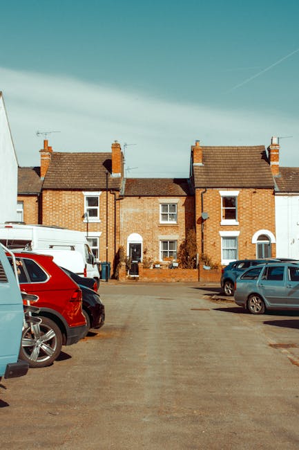 A row of three connected brick houses with pitched roofs and chimneys, situated directly behind a parking area with several parked vehicles including a red SUV, a white van, and a grey car. The pavement and parking lot are visible in the foreground, with a clear sky overhead. The photograph captures an exterior scene during daylight, likely part of a house removal or relocation process, with the company Epsom Man and Van providing removals services as indicated by the context of the KT18 parking and access guide for Epsom house removals.