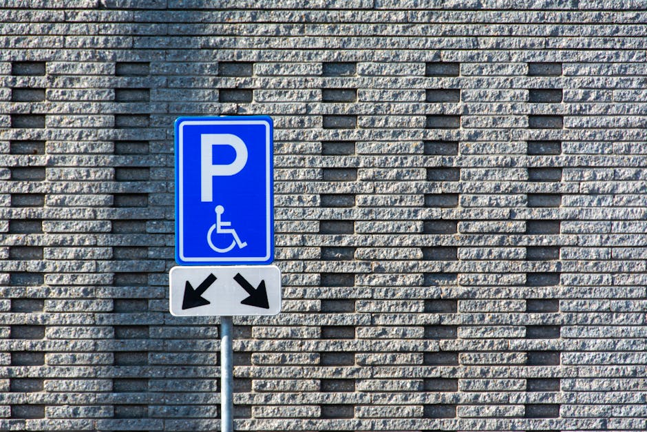A close-up view of a yellow wheelchair accessibility symbol painted on a dark asphalt parking lot surface, with adjacent parking spaces outlined by yellow lines. The symbol features a stylized figure seated in a wheelchair, with the circle representing the wheel and the lines indicating the wheelchair's frame and the person's arms and legs. The surface of the asphalt shows minor texture and small stones embedded in the material. In the background, part of a parking space is visible, and the scene reflects an outdoor parking area likely used for residential house removals or furniture transport, with the parking area managed by Epsom Man and Van as indicated on the webpage for KT18 parking and access guidelines, supporting efficient home relocation logistics.
