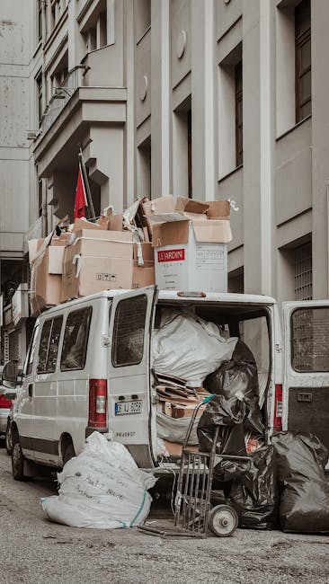 A white commercial van parked on a city street beside a multi-storey building with large, beige façade walls and multiple windows. The van's rear doors are open, revealing a load of various packing materials, including large white plastic bags, cardboard boxes, and wrapped items, indicating a home relocation or furniture transport process. Several cardboard boxes, some open and others sealed, are stacked on top of the van and on the pavement nearby. A small trolley and black garbage bags are positioned on the ground in front of the van, while the packaging materials inside the van include plastic wrap and foam padding. The scene is well-lit with natural daylight, capturing the typical logistical setup involved in house removals, with a focus on loading and packing efforts associated with professional moving services like those provided by Epsom Man and Van.