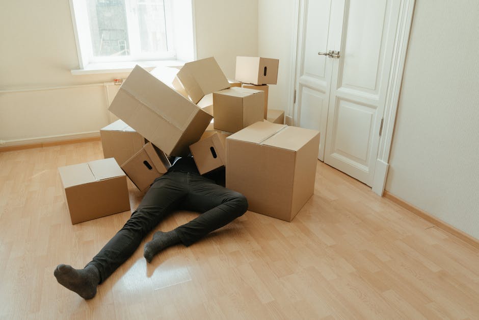 A person lying on the light wooden floor of a room, partially obscured by stacked cardboard moving boxes in various sizes, some positioned upright and others on their sides. The individual is wearing dark pants and grey socks, with their legs extended and one arm partially visible under the boxes. The room has cream-colored walls, a white door with a silver handle, and a window nearby allowing natural light to illuminate the space. The office or living area appears prepared for a home relocation, with boxes likely packed for a house or flat move. The scene illustrates the packing phase of furniture transport and moving logistics, and the presence of damage or access issues can be inferred from the disorganized placement of boxes and the person's position, possibly indicating difficulties during a move. Epsom Man and Van, a professional removals service, may be involved in addressing such packing problems and access challenges during house removals.