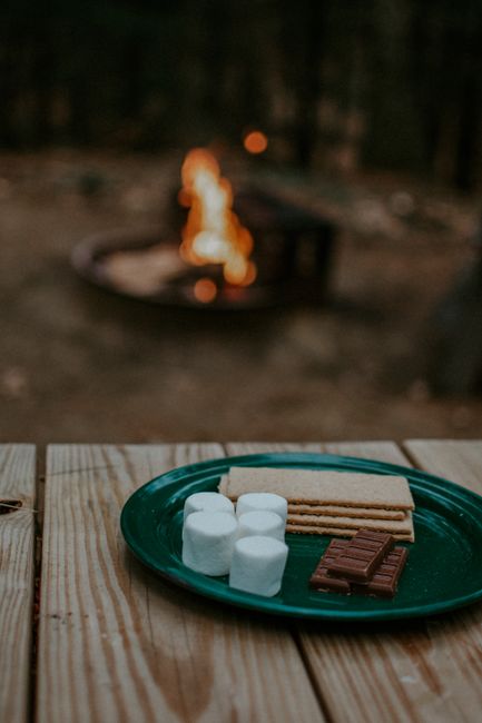 A green plastic plate on a wooden outdoor surface holds several marshmallows, some rectangular chocolate bars, and pieces of graham crackers, arranged for making s'mores. The background features a blurred outdoor scene with a campfire burning in a metal fire pit, emitting flames and small glowing embers, suggesting an outdoor camping or recreational setting. The natural light and the warm glow from the fire highlight the casual and relaxed environment. This image is associated with home relocation or outdoor activities involving packing and moving essentials, as seen in the context of Epsom Man and Van's removals services, though the focus remains on the outdoor snack setup.